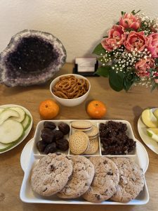 Auf einem Tisch stehen verschiedene Snacks und aufgeschnittene Birnen und Äpfel, im Hintergrund rechts ein Strauss mit Tulpen und links eine große Amethyst-Druse