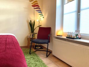 Blick in einen Massageraum, links eine Liege mit dunkelroter Decke, in der rechten Ecke Stuhl, Pflanze und Lampe, rechts ein Fenster mit Sichtschutz.