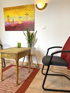 Sitzgruppe mit Metallstuhl und kleinem Holztisch, an der Wand ein gemaltes Bild mit tanzenden Frauen, darunter eine Pflanze.